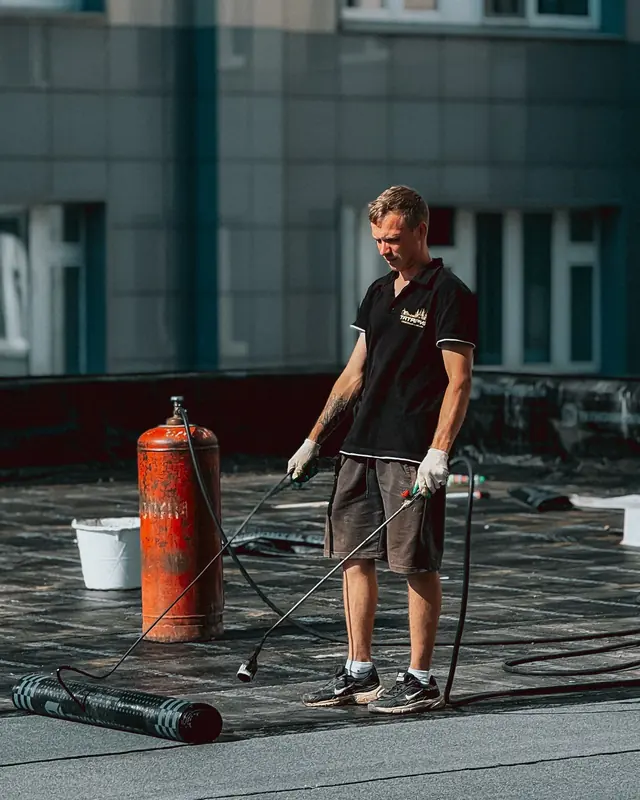 A roofer with a gas burner standing beside a membrane roll and gas cylinder on a flat roof